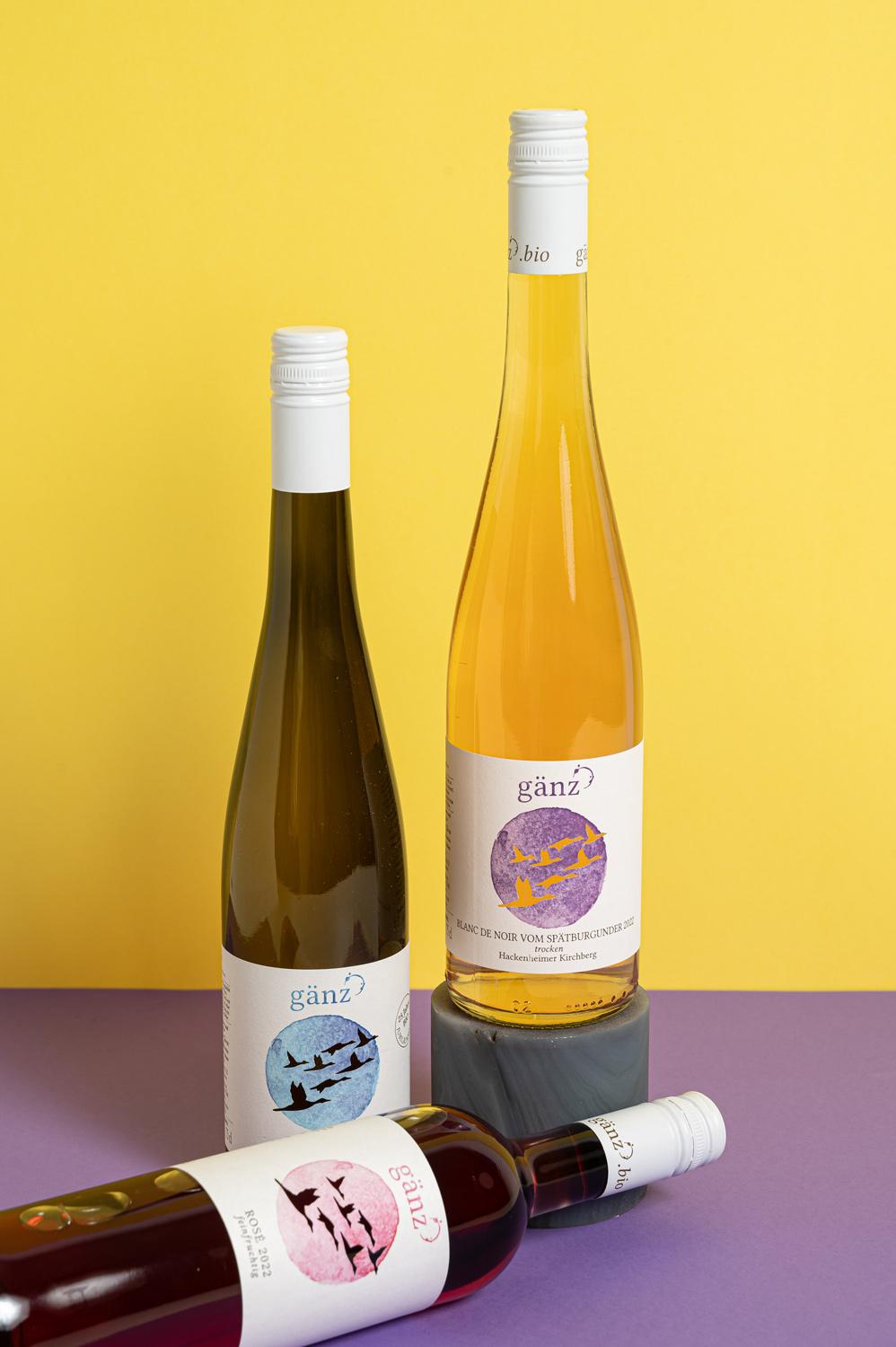 Drei Weinflaschen auf einem lila Untergrund und gelben Hintergrund.