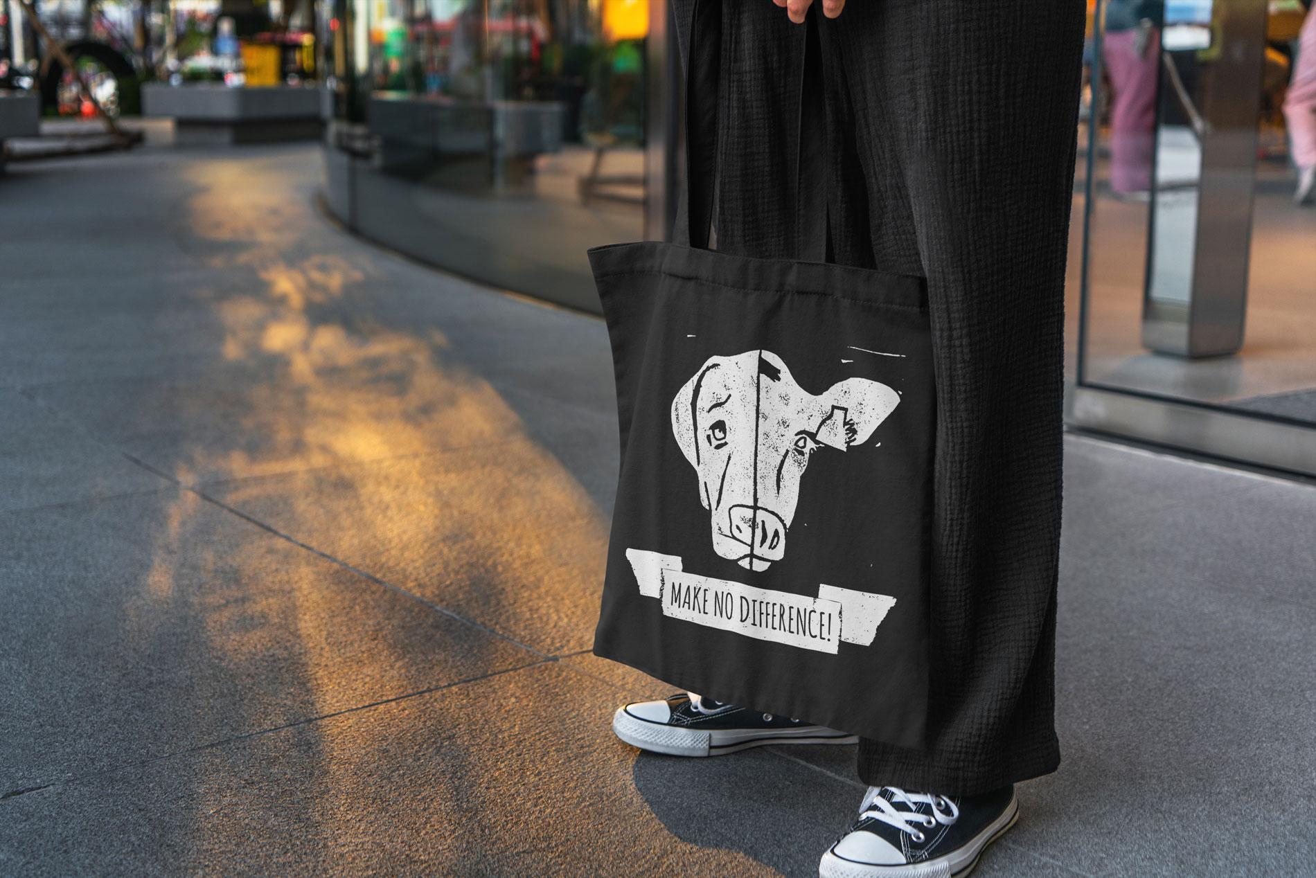 Person trägt eine schwarze Baumwolltasche mit einem weißen Print einer Kuh und dem Schriftzug 'Make No Difference!' Die Tasche vermittelt eine klare vegane Botschaft. Die Person trägt lässige schwarze Kleidung und Converse-Sneaker, während sie auf einer urbanen Straße steht. Der Hintergrund zeigt eine moderne Glasfassade, beleuchtet von warmem Licht, was der Szene eine städtische und zeitgemäße Atmosphäre verleiht.